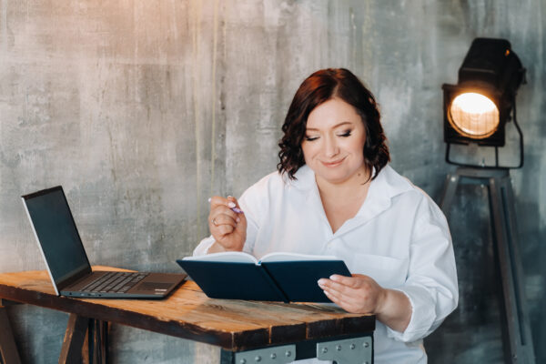A Businesswoman In A White Shirt Sits At A Desk Writing Something In A Notebook In The Office